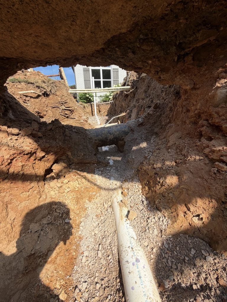 Excavated trench exposing a broken sewer line beside a home